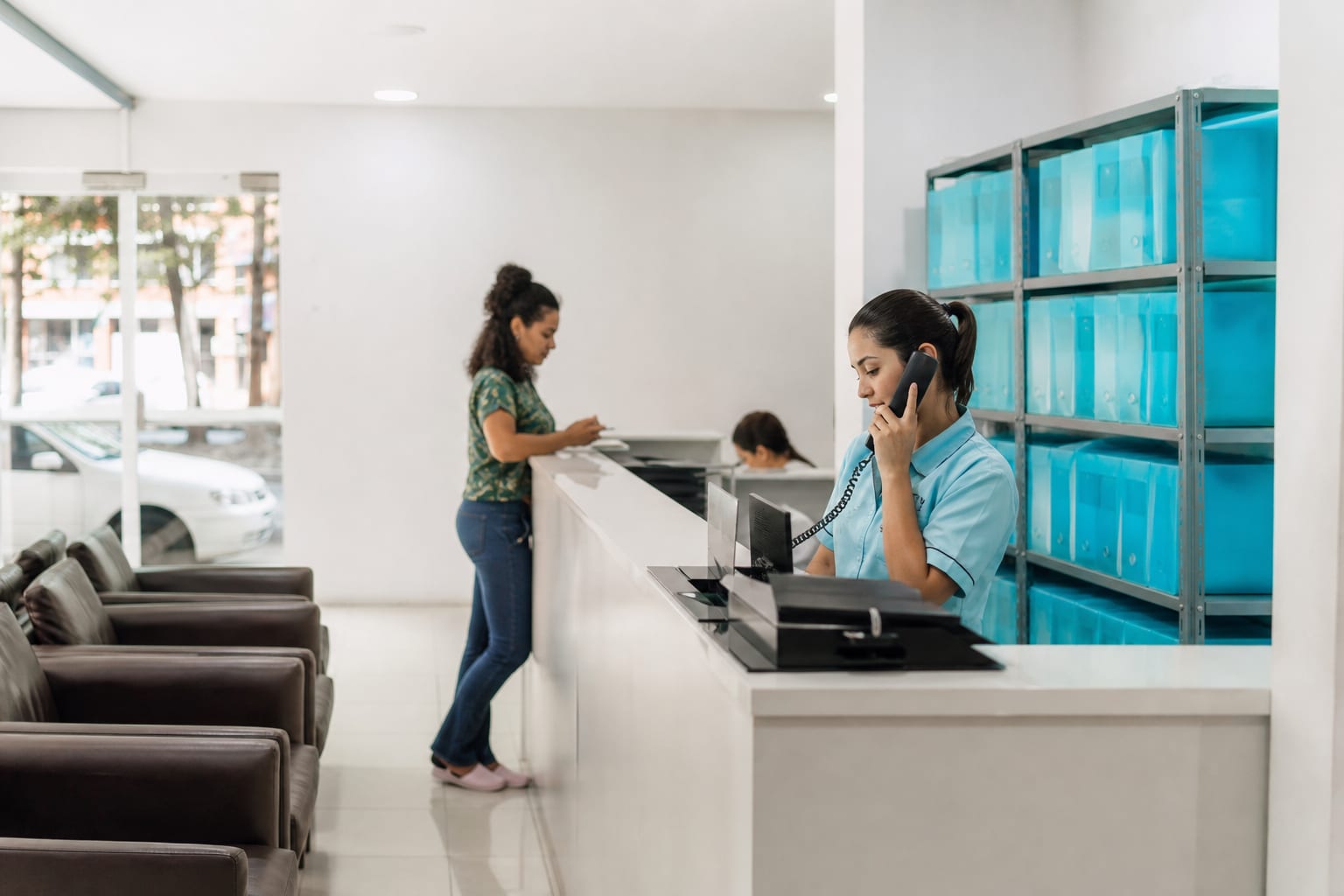 Recepcionista atendendo ao telefone no balcão de uma pequena clínica, com poltronas de espera ao lado