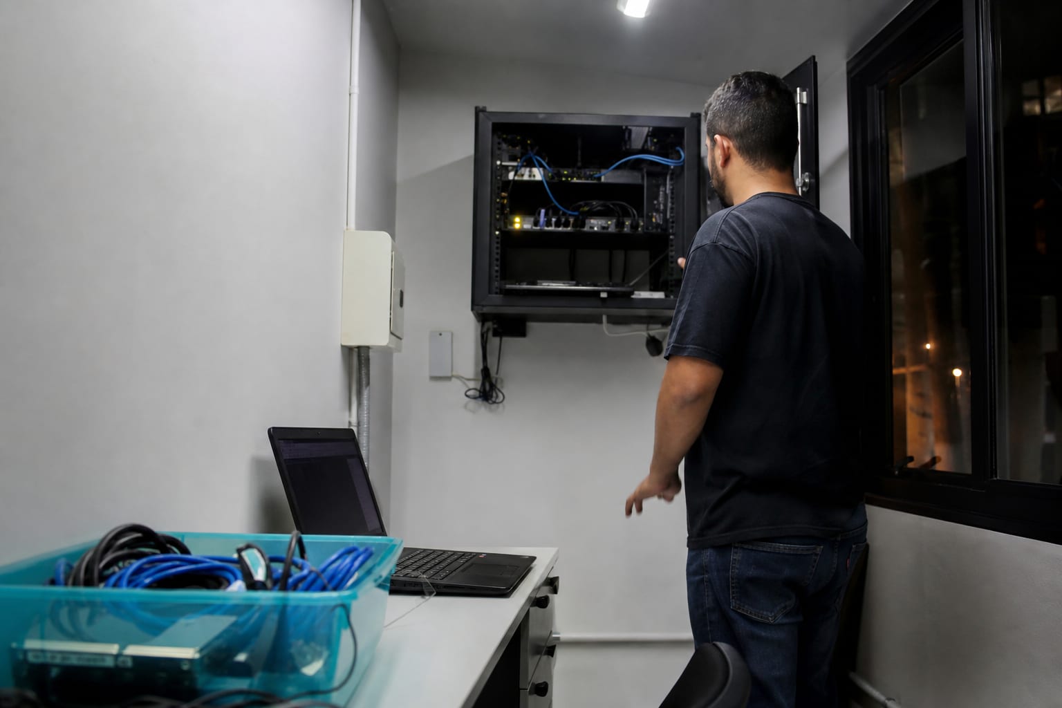 Técnico com notebook em pé diante de um rack de rede aberto com cabos azuis organizados